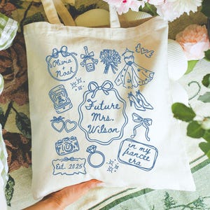 May include: Cream-colored tote bag with blue illustrations and text. The bag features drawings of wedding-related items and the text "Future Mrs. Wilson" and "in my fiancee era". The bag also has the names "Olivia Noah" and the year "Est. 2025".