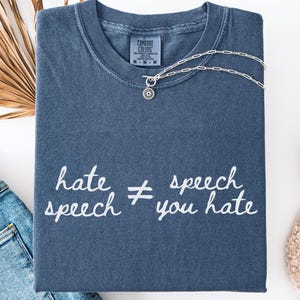 May include: A denim blue t-shirt with the text "hate speech &ne; you hate" in white script. A silver necklace with a small pendant rests on the shirt. The shirt is folded neatly, with a pair of jeans and a white shoe visible.