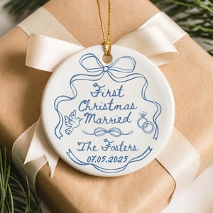 May include: A white ceramic Christmas ornament with blue text and design, including the words "First Christmas Married," a dove, and wedding rings. The ornament hangs from a gold string and rests on a wrapped gift with a cream ribbon.