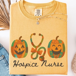 May include: Mustard-colored t-shirt with a Halloween-themed design. The graphic includes two jack-o'-lanterns flanking a stethoscope, with the words "Hospice Nurse" below. The shirt is folded, with a necklace and jeans visible.