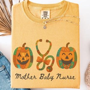 May include: Mustard-colored t-shirt with a pumpkin and stethoscope graphic. The shirt features two smiling jack-o'-lanterns flanking a stethoscope, with the words "Mother Baby Nurse" below. A gold necklace and jeans are next to the shirt.