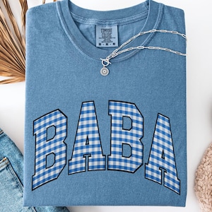 Peut inclure: T-shirt bleu jean avec le mot "BABA" en motif vichy bleu et blanc. Le t-shirt est plié et accessoirisé d'un collier argenté. Un jean bleu et une chaussure blanche sont également visibles.