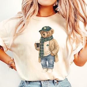 May include: Ivory-colored t-shirt featuring a cartoon bear dressed in a green cap, scarf, and coat, holding a pint of dark beer. The bear wears blue jeans and white sneakers. The shirt has the word "IVORY" at the bottom.