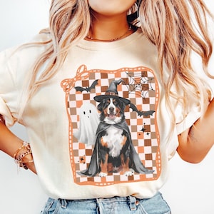 May include: Ivory-colored t-shirt featuring a Halloween-themed graphic. The graphic includes a Bernese Mountain Dog dressed as a witch, a ghost, bats, and a spiderweb, set against a checkered background. The word "IVORY" is printed at the bottom.