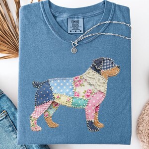 Peut inclure: T-shirt bleu jean avec un motif de chien en patchwork. Le chien est composé de divers motifs de tissu, dont des fleurs et des pois. Un collier en argent avec un pendentif repose sur le t-shirt. Le t-shirt est plié avec un jean et une chaussure.