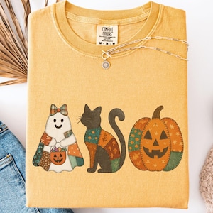 May include: Mustard-colored t-shirt with a Halloween-themed graphic. The graphic features a ghost, a black cat, and a jack-o'-lantern. The shirt is folded and displayed with a pair of blue jeans, a necklace, and a pair of white shoes.