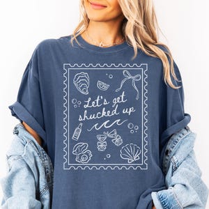 May include: A denim blue t-shirt with a white graphic that reads "Let's get shucked up" inside a stamp-like border. The design includes illustrations of oysters, a bottle, and seashells. The shirt is a relaxed fit.