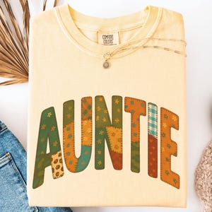 May include: A light yellow sweatshirt with the word "AUNTIE" in a patchwork design. The sweatshirt is folded and has a Comfort Colors tag. A gold necklace and a pair of blue jeans are also visible.
