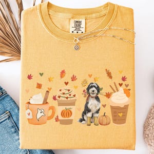 May include: Mustard-colored Comfort Colors t-shirt with a fall-themed graphic. The graphic features coffee cups, autumn leaves, hearts, and a dog wearing a scarf and boots. A gold necklace is draped across the shirt. The shirt is folded on a white surface.