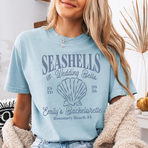 May include: Light blue t-shirt with the text "Seashells & Wedding Bells" and a seashell graphic. The text "Emily's Bachelorette Rosemary Beach, FL" is also on the shirt.