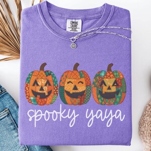 May include: A violet Comfort Colors sweatshirt with three smiling jack-o'-lanterns in patchwork designs. The words "spooky yaya" are printed below the pumpkins. A silver necklace with a pendant is draped on the shirt.