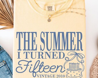 May include: A yellow t-shirt with the text "THE SUMMER I TURNED Fifteen VINTAGE 2010" in blue. The design includes a cake with candles. The shirt is paired with blue jeans, a necklace, and white sneakers.
