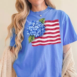 Op de afbeelding: Een korenbloemblauw T-shirt met korte mouwen en een grafische print van een blauwe hortensiabloem die een gestileerde Amerikaanse vlag met rode en witte horizontale strepen overlapt. Het shirt heeft een ronde hals en een ruime pasvorm.