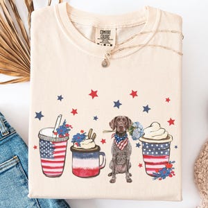 May include: Ivory t-shirt featuring a graphic of a dog holding a bouquet of flowers, a red, white and blue cup of coffee, a red, white and blue cup of ice cream, and a red, white and blue cup of soda. The graphic is surrounded by red and blue stars.