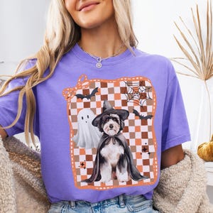 May include: A violet t-shirt featuring a Halloween-themed graphic. The graphic includes a dog in a witch hat and cape, a ghost, bats, and a spiderweb on a checkered background. The word "VIOLET" is at the bottom.