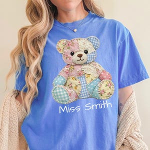 May include: A blue t-shirt with a patchwork teddy bear design in floral and gingham patterns. The text "Miss Smith" is printed below the bear. The shirt has a relaxed fit.