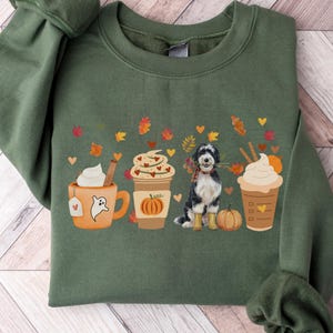 May include: Military green sweatshirt featuring a fall-themed graphic. The design includes a dog, pumpkins, and coffee cups with whipped cream and cinnamon sticks. The sweatshirt has long sleeves and a crew neck.