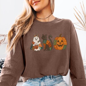 May include: Espresso-colored sweatshirt featuring a Halloween-themed design. The graphic includes a ghost, a black cat, and a jack-o'-lantern. The ghost and cat are adorned with patchwork patterns. The sweatshirt is a long-sleeved crew neck.