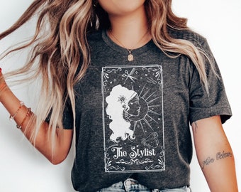 May include: A dark grey heather t-shirt with a white graphic of a tarot card design. The card features a stylised illustration of a woman with a sun and moon design. The text "The Stylist" is printed on the card.