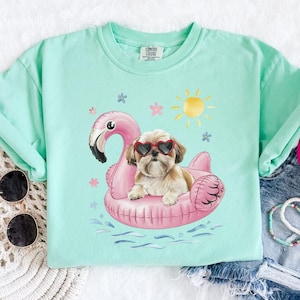 May include: A light green t-shirt with a graphic of a dog wearing sunglasses and riding a pink flamingo pool float. The dog is a Shih Tzu breed. The graphic is surrounded by small pink flowers and a yellow sun.