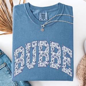 May include: Blue jean colored t-shirt with the word "BUBBE" in a floral pattern. The shirt is folded and has a silver necklace with a pendant. A pair of blue jeans and a white shoe are also visible.