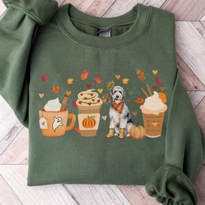May include: Military green sweatshirt featuring a fall-themed graphic. The design includes coffee cups, a dog wearing a scarf and boots, pumpkins, and autumn leaves. The text "MILITARY GREEN" is at the bottom of the image.