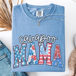 May include: A light blue t-shirt with the words "cavapoo MAMA" in a patriotic design. The letters are filled with stars and stripes. A silver necklace with a pendant is on the shirt. The shirt is folded next to a pair of jeans.