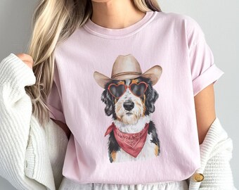 May include: A pink t-shirt with a graphic of a dog wearing a cowboy hat and heart-shaped sunglasses. The dog is wearing a red bandana around its neck.