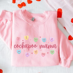 May include: Pink sweatshirt with the text "cockapoo mama" and a design of candy hearts with phrases like "puppy love", "I die you", "must love dogs", "I chew you", "love bites", "xoxo", "treat sealed", "I woof you", "pawsitive vibes", "fur-ever mine".