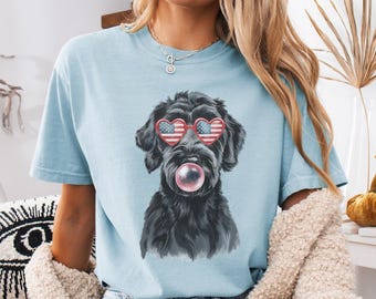 Puede incluir: Una camiseta azul claro con un perro negro que lleva gafas de sol en forma de corazón rojo, blanco y azul y sopla una burbuja. El texto "CHAMBRAY" es visible en la parte inferior de la imagen.