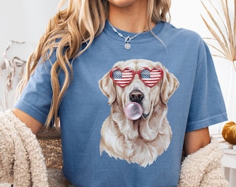 Puede incluir: Una camiseta azul vaquero con un gráfico de un golden retriever que lleva gafas de corazón rojo, blanco y azul y mastica chicle. El perro está mirando directamente al espectador.