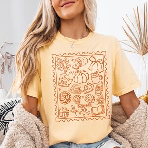 May include: Butter-colored t-shirt featuring a brown stamp-like design with fall-themed illustrations. The design includes a pumpkin, teapot, cupcakes, and a book. The word "BUTTER" is printed at the bottom of the image.