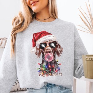 May include: A light gray sweatshirt featuring a graphic of a dog wearing sunglasses and a Santa hat, blowing a bubblegum bubble. The dog is adorned with Christmas lights. The word "ASH" is printed at the bottom.