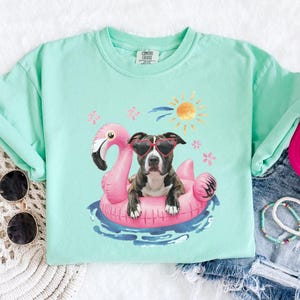 May include: A light green t-shirt featuring a graphic of a dog wearing sunglasses and floating on a pink flamingo pool float. The dog is a brindle pit bull mix. The graphic is surrounded by pink flowers and a yellow sun.