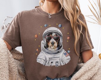 May include: A brown t-shirt featuring a graphic of a dog wearing an astronaut suit and heart-shaped sunglasses. The dog is surrounded by stars and planets. The text "ESPRESSO" is visible at the bottom of the image.