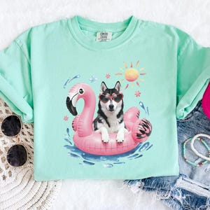 May include: A light green t-shirt featuring a graphic of a black and white husky dog wearing heart-shaped sunglasses and riding a pink flamingo pool float. The dog is in a pool of blue water with a yellow sun and white flowers in the background.