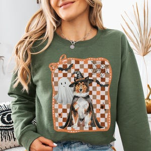 May include: Military green sweatshirt featuring a Halloween-themed graphic. The graphic includes a dog in a witch's hat and cape, a ghost, bats, and a spiderweb on a checkered background. The sweatshirt is a crew neck style.