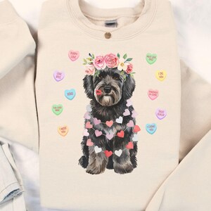 May include: A sand-colored sweatshirt featuring a watercolor illustration of a black dog adorned with a floral crown and a garland of hearts. Candy hearts with phrases like "Puppy Love" and "I Like You" are scattered around the dog.