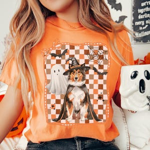 May include: Orange t-shirt with a Halloween-themed graphic featuring a dog in a witch costume, a ghost, bats, and a spiderweb. The shirt has a checkered background and the word "MELON" at the bottom.