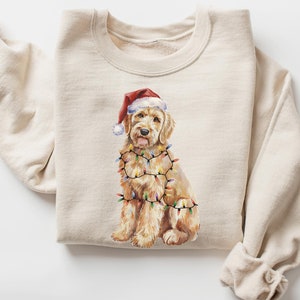 May include: A beige sweatshirt featuring a Christmas-themed illustration of a golden doodle dog wearing a Santa hat and tangled in Christmas lights.