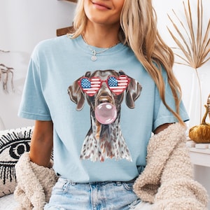 May include: A light blue t-shirt with a graphic of a brown and white dog wearing red, white, and blue heart-shaped sunglasses and blowing a pink bubble. The text "CHAMBRAY" is visible at the bottom of the image.