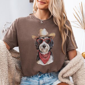 May include: A brown t-shirt with a graphic of a dog wearing a cowboy hat and heart-shaped sunglasses. The dog is wearing a red bandana around its neck.