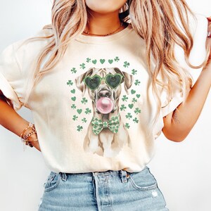 Peut inclure: Un t-shirt ivoire clair avec un motif représentant un chien portant des lunettes de soleil en forme de cœur vertes et un nœud papillon vert. Le chien tient une bulle rose dans sa gueule. Le motif est entouré de trèfles et de cœurs verts.