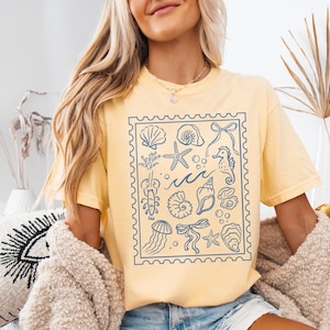 May include: Butter-colored t-shirt featuring a blue stamp design with various sea creatures and elements, including seashells, starfish, and seahorses. The word "BUTTER" is at the bottom of the image.