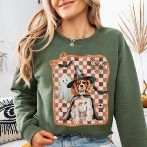 May include: Military green sweatshirt with a Halloween graphic. The design features a Cavalier King Charles Spaniel in a witch hat and cape, a ghost, bats, and a spiderweb on a checkered background.