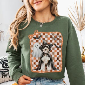 May include: Military green sweatshirt featuring a Halloween-themed graphic. The graphic includes a dog dressed as a witch, a ghost, bats, and a spiderweb on a checkered background. The sweatshirt is a crew neck style.