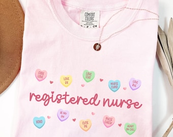 以下が含まれることがあります： 「registered nurse」という文字と、「Love Stat」、「Nurse Cupid」、「XOXO」などのフレーズが書かれたキャンディーハートのグラフィックがプリントされたピンクのTシャツ。