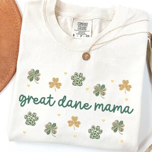 Peut inclure: T-shirt ivoire avec un motif vert et or qui dit "great dane mama". Le motif comprend des empreintes de pattes et des trèfles.