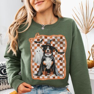 May include: Military green sweatshirt featuring a Halloween-themed graphic. The graphic includes a Bernese Mountain Dog dressed as a witch, a ghost, bats, and a spiderweb, set against a checkered background. The sweatshirt is a crew neck style.