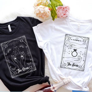 May include: Two t-shirts, one black and one white, with a graphic design of hands holding a ring and celestial symbols. The text on the black shirt reads 'The Groom' and the text on the white shirt reads 'The Bride.'